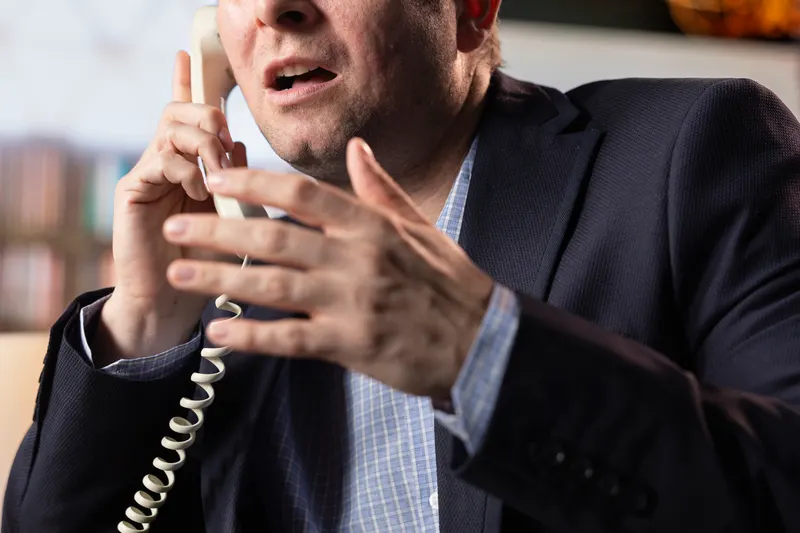 A man mid-conversation on a corded desk phone, gesturing with one hand
